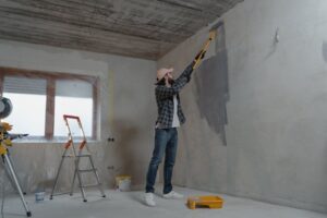 גבר מצייר קיר במהלך שיפוץ חדר עם רולר ארוך, סולם וכלי צבע.